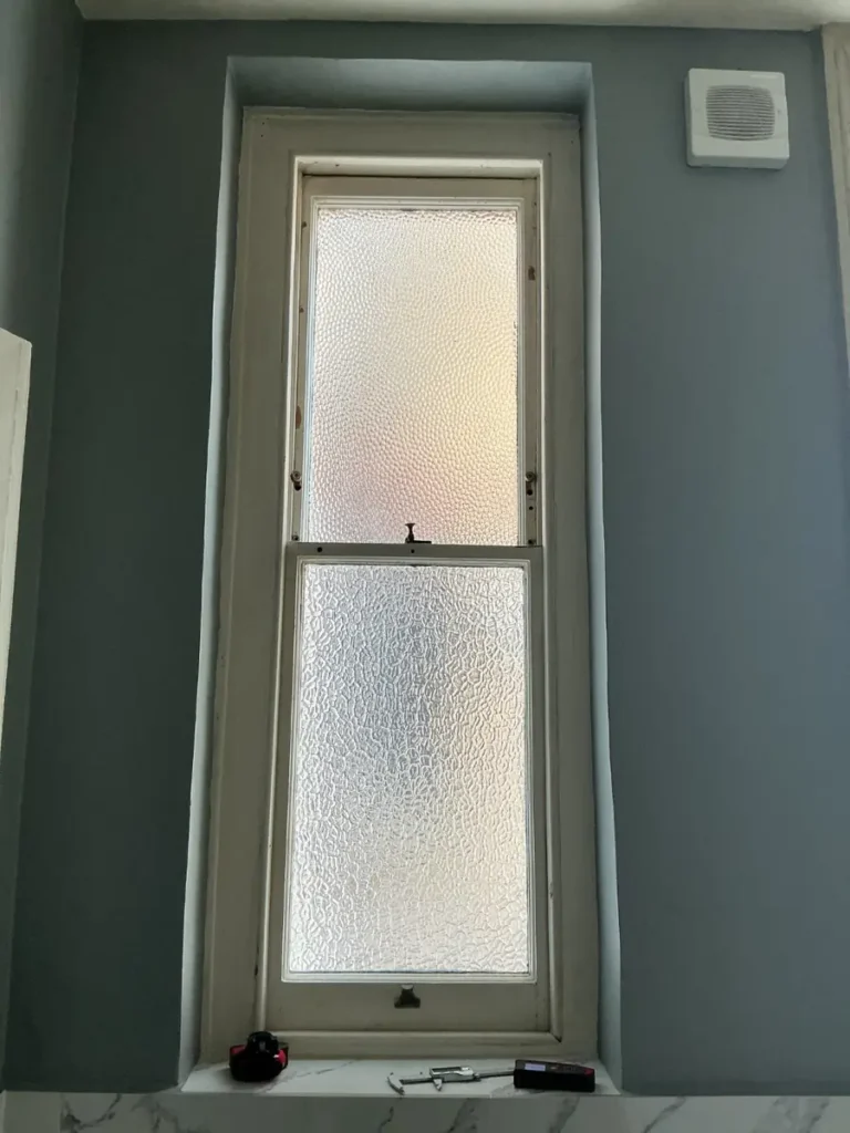 Original sash window with obscured glass above a marble-tiled cill in a Battersea bathroom